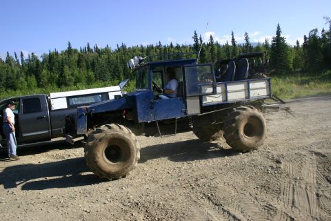 2003 Jeep TJ - A heavily modified blue truck with oversized tires sits on a dirt road beside a pickup truck.