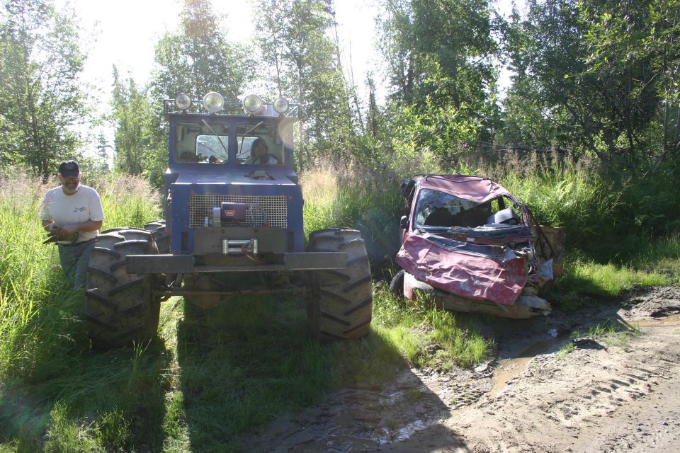 Wrecked red car with crumpled front end and broken windshield, parked on a muddy dirt road next to a large blue vehicle
