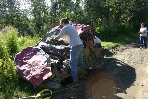 2003 Jeep TJ - A severely damaged red car sits off a dirt road, with two people observing the scene.