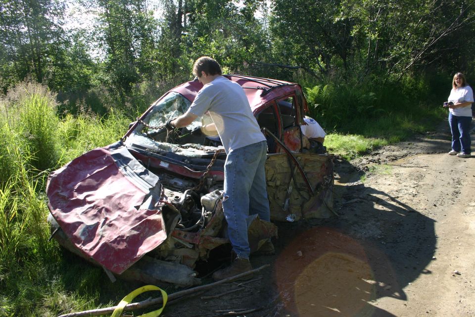 Damaged red car with crumpled front end, broken windshield, and exposed engine parts, off a dirt road