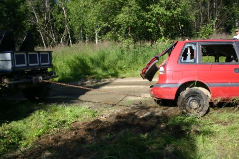2003 Jeep TJ - A red SUV is being towed by a truck on a dirt road, with its rear hatch open and window broken.