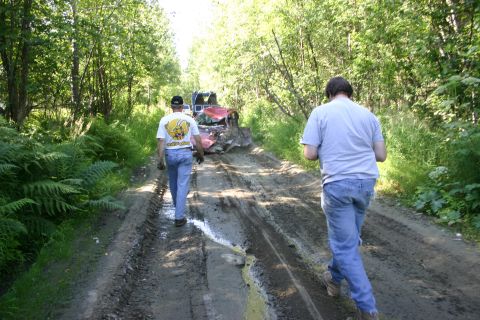2003 Jeep TJ - A muddy track winds through trees, with two men walking towards a damaged vehicle and a truck.