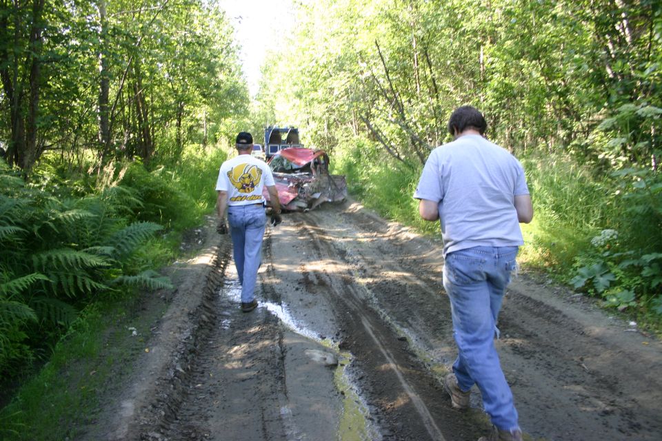 Two guys walking down a muddy path toward a wrecked red car and a blue truck