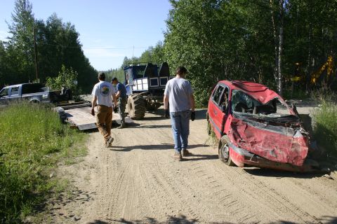 2003 Jeep TJ - A severely damaged red car sits on a dirt road next to a large industrial vehicle and a pickup truck.