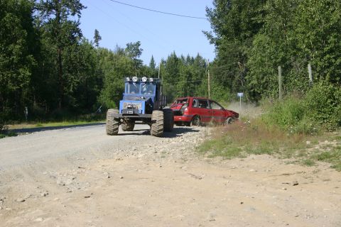 2003 Jeep TJ - A blue tractor tows a red car that appears to be damaged and is kicking up dust on a rural dirt road.
