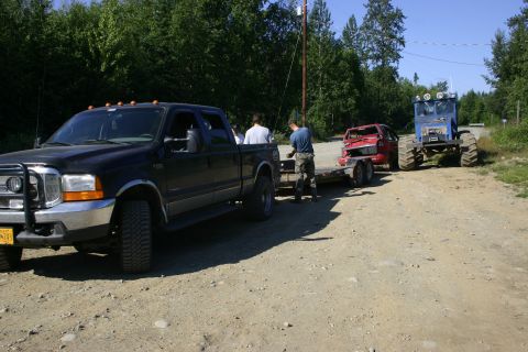 2003 Jeep TJ - Black pickup truck tows a damaged red car on a trailer, followed by a large blue tractor.
