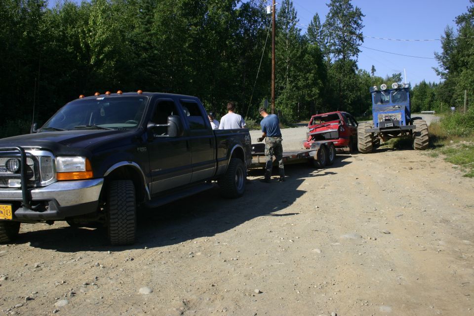 Black Ford F-Series pickup towing a red car on a trailer, with a blue tractor behind it on a dirt road