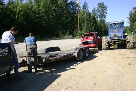2003 Jeep TJ - A damaged red car is loaded onto a trailer, with a large blue vehicle positioned behind it.