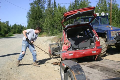2003 Jeep TJ - A person is using a metal bar to help pull a red SUV onto a trailer, with a large blue tractor in the background.