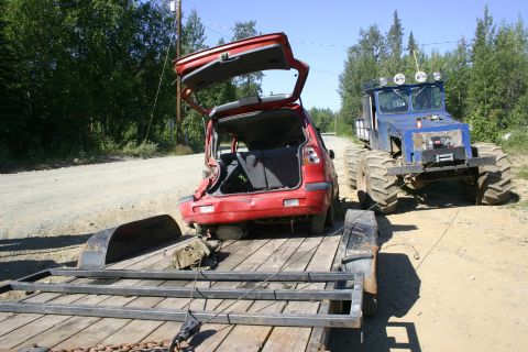 2003 Jeep TJ - A red car is loaded onto a trailer and being towed by a blue all-terrain vehicle with oversized tires.