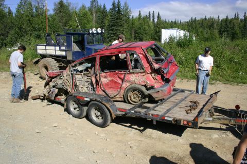 2003 Jeep TJ - A heavily damaged red car is loaded onto a trailer, with a large blue truck nearby.