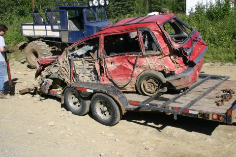 2003 Jeep TJ - A heavily damaged red car is loaded onto a trailer, with a large blue truck visible behind it.