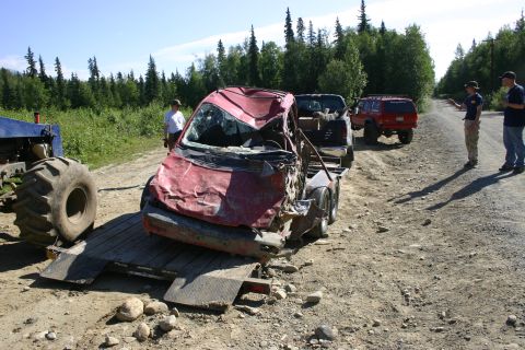 2003 Jeep TJ - A severely damaged red vehicle is loaded onto a trailer, with other vehicles and people present on a dirt road.