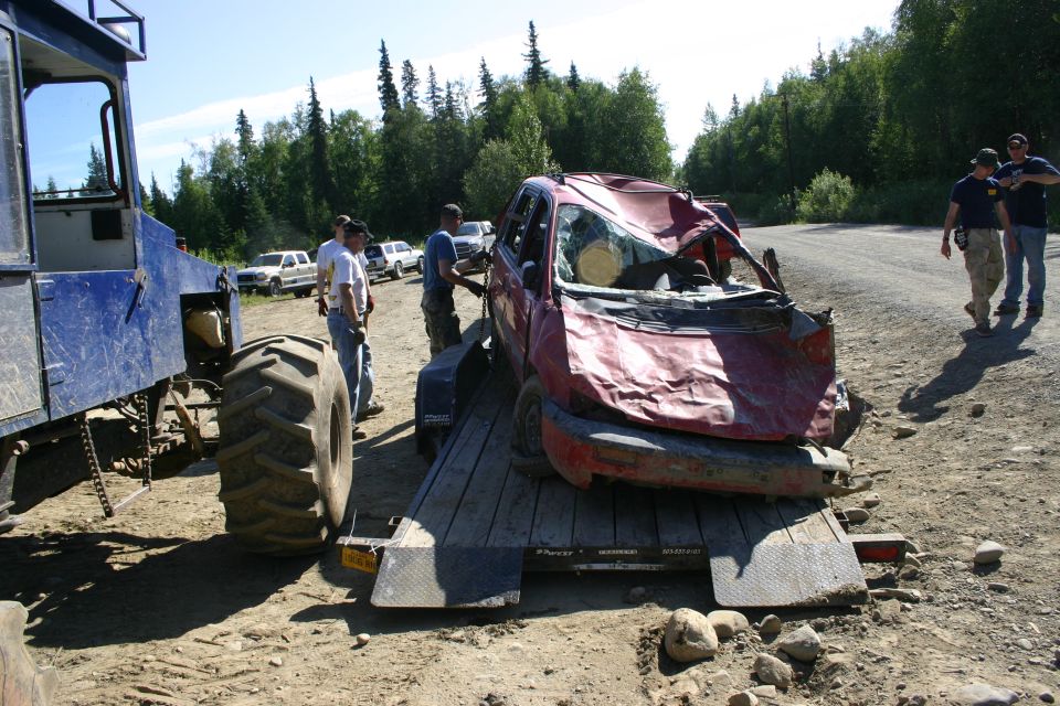 Damaged red car on a flatbed trailer, front end crushed, windshield broken. Blue tractor unit to the left