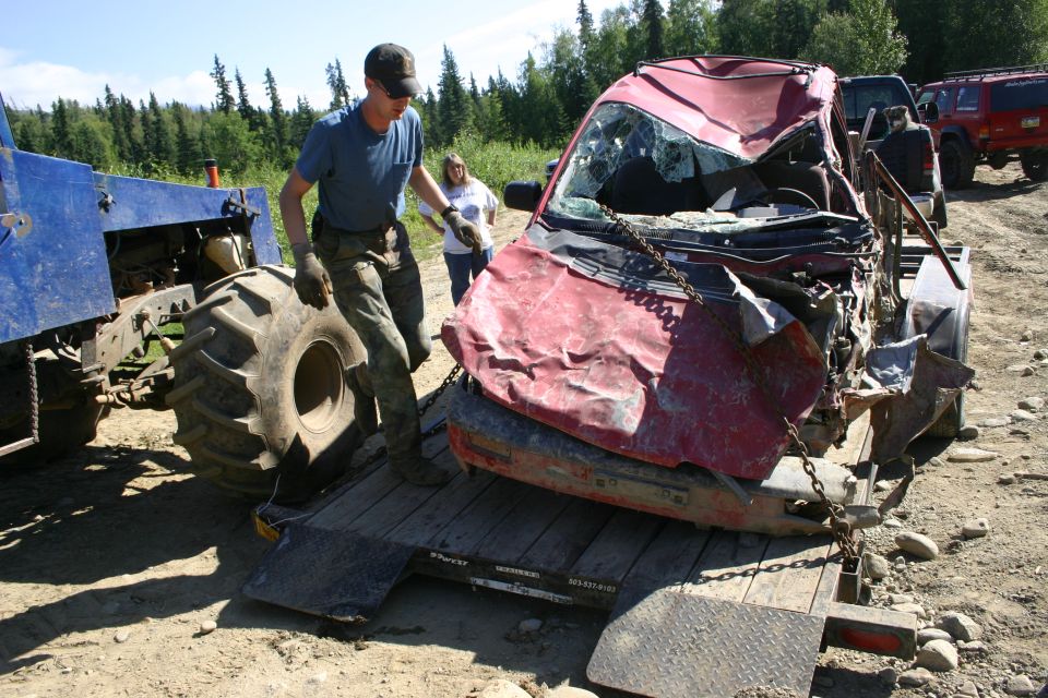 Man loading a heavily damaged red SUV onto a flatbed trailer with a blue tractor. Another SUV is parked behind
