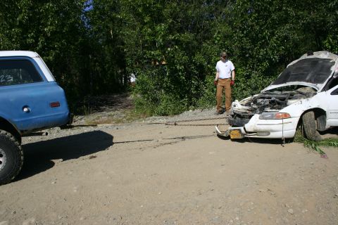 2003 Jeep TJ - A white sedan, its hood open and front end damaged, is being towed by a blue SUV on a dirt road.