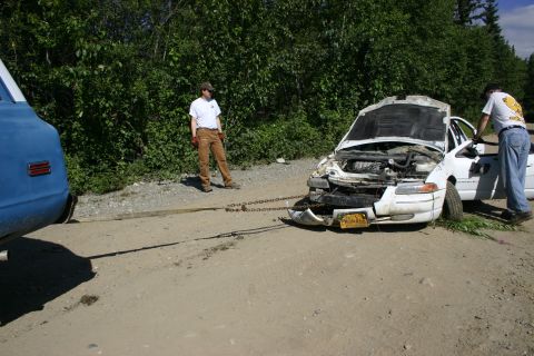 2003 Jeep TJ - A damaged white sedan is being towed by a blue truck on a dirt road.
