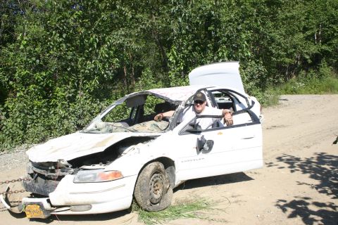 2003 Jeep TJ - A heavily damaged white sedan sits on a dirt road, with a person visible inside the gutted interior.