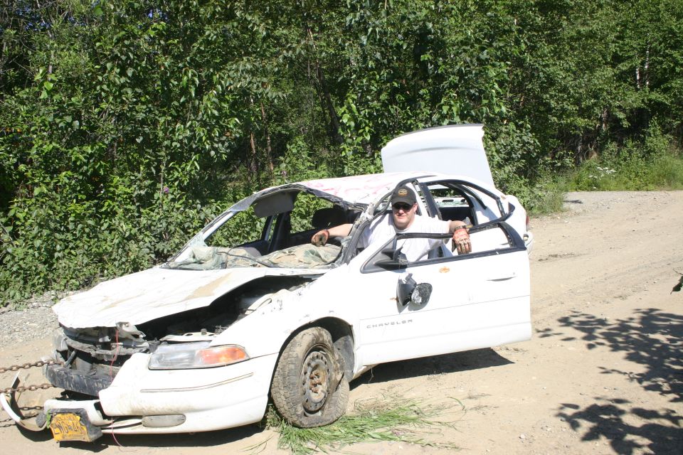 White sedan with severe front-end damage, roof cut open, and a person sitting in the driver's seat. The word 'CHRYSLER' is visible on the door