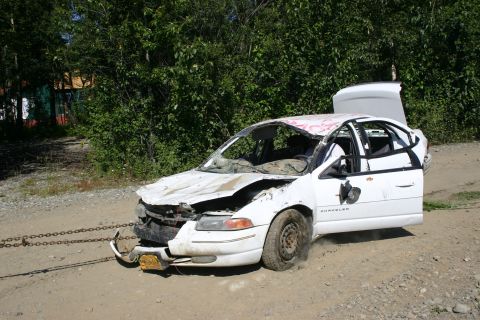 2003 Jeep TJ - A heavily damaged white sedan sits on a dirt road, its trunk and driver's door open, with chains attached to the front bumper.
