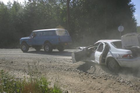 2003 Jeep TJ - A blue SUV tows a damaged white sedan on a dusty dirt road.