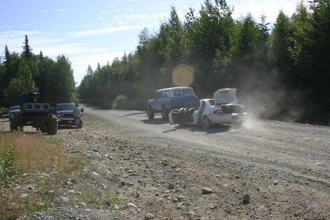 2003 Jeep TJ - A group of vehicles, including a heavily modified truck and an SUV towing a damaged car, travel on a remote dirt road.