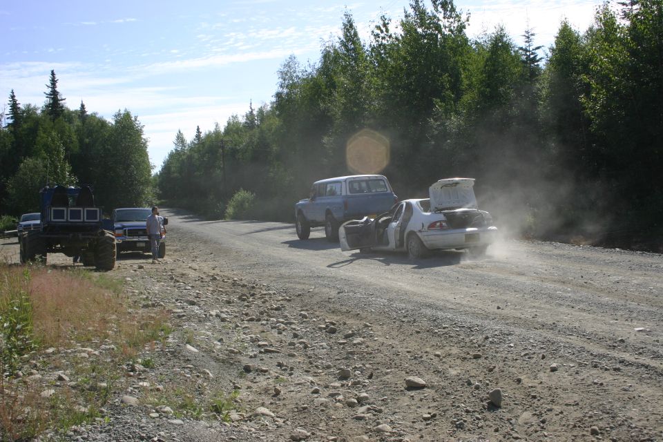 Several vehicles, including a large off-road truck and a blue SUV towing a white car with its trunk open, are on a dusty dirt road