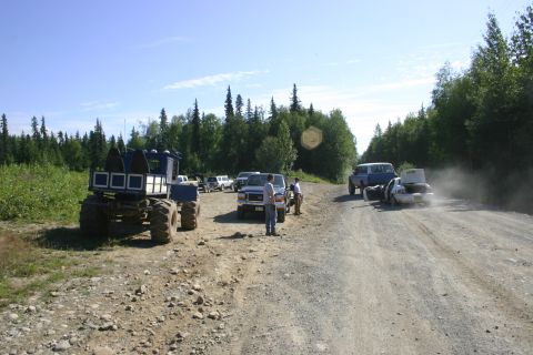2003 Jeep TJ - Several vehicles are gathered on a dusty dirt road surrounded by trees undergrowth and trees under a clear blue sky.