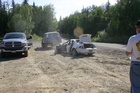 2003 Jeep TJ - A white sedan, stripped of its doors and with its trunk open, is towed by a blue SUV on a dirt road, with a pickup truck and trailer nearby.
