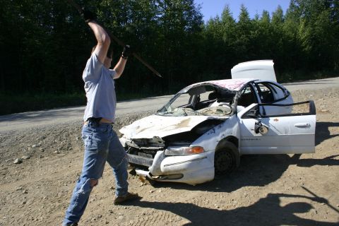 2003 Jeep TJ - A person prepares to strike a severely damaged white sedan with a metal pole in an outdoor setting.