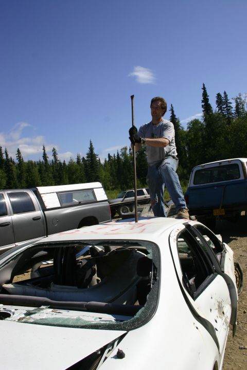 2003 Jeep TJ - A man stands on the roof of a damaged white sedan in a junkyard, with other vehicles and trees visible.