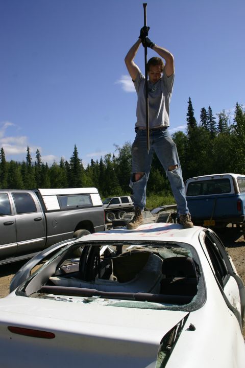 2003 Jeep TJ - A man stands on the roof of a damaged white car, holding a crowbar, with other vehicles visible in the background.