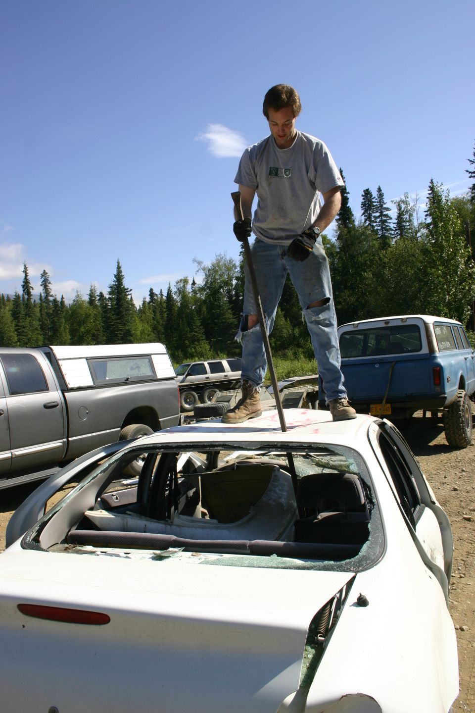 Man with crowbar on wrecked white car roof, gray pickup and blue truck in background