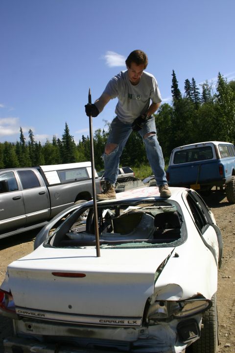 2003 Jeep TJ - A man stands on the roof of a heavily damaged white sedan in a junkyard setting.
