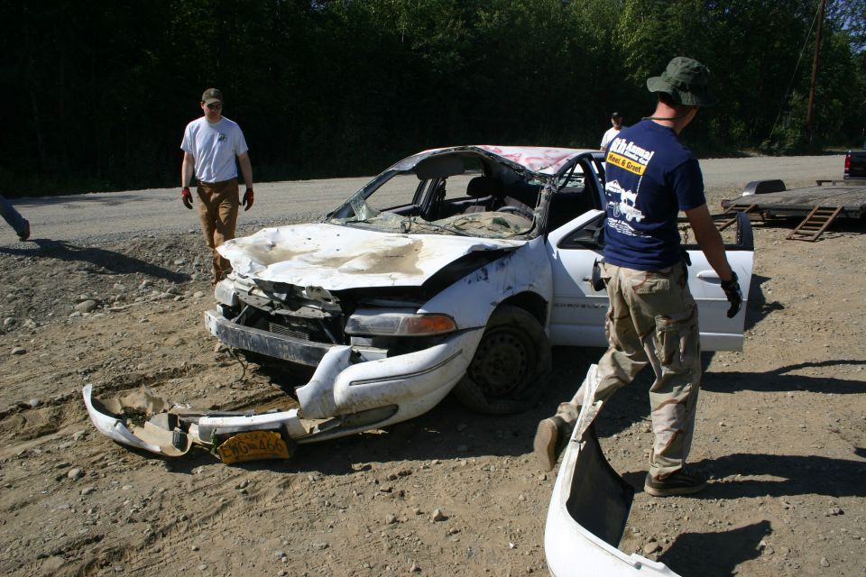 Wrecked white sedan with front bumper detached and hood crumpled. Two men stand nearby