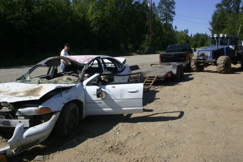 2003 Jeep TJ - A heavily damaged white sedan is being towed by a blue tractor, with a black pickup truck also visible.