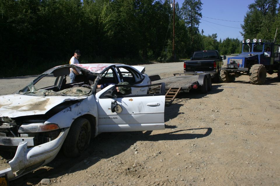 Damaged white sedan with open driver door, red graffiti on roof, being towed by a blue tractor