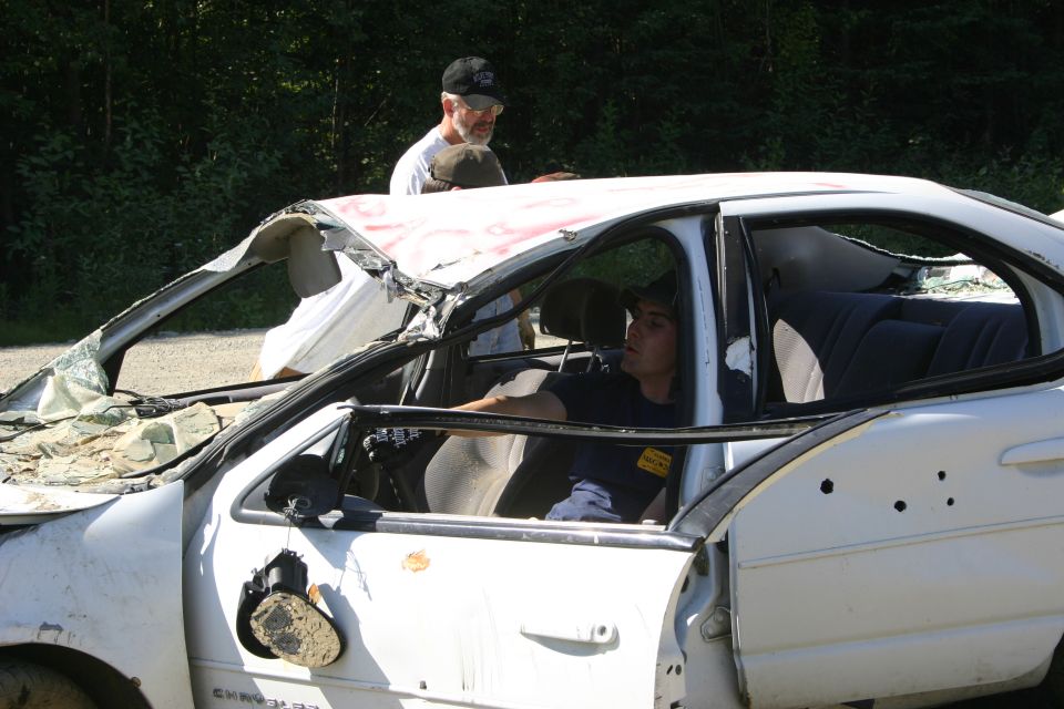 White sedan with severe roof and door damage, two men visible inside and near the vehicle