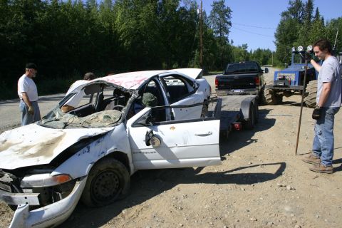 2003 Jeep TJ - A heavily damaged white sedan is being towed, with a black pickup truck and a blue tractor visible in the background.