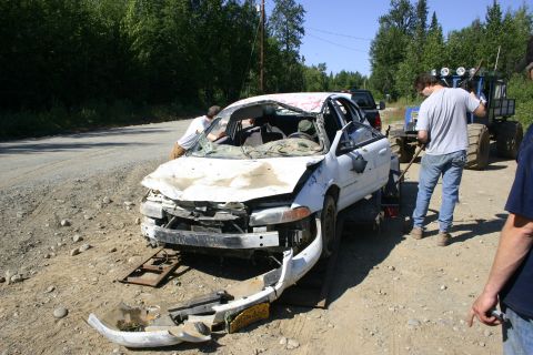 2003 Jeep TJ - Wrecked white sedan being towed with a tractor assisting. The front end is heavily damaged.