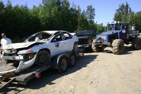 2003 Jeep TJ - A heavily damaged white sedan is transported on a trailer, pulled by a custom blue off-road vehicle.