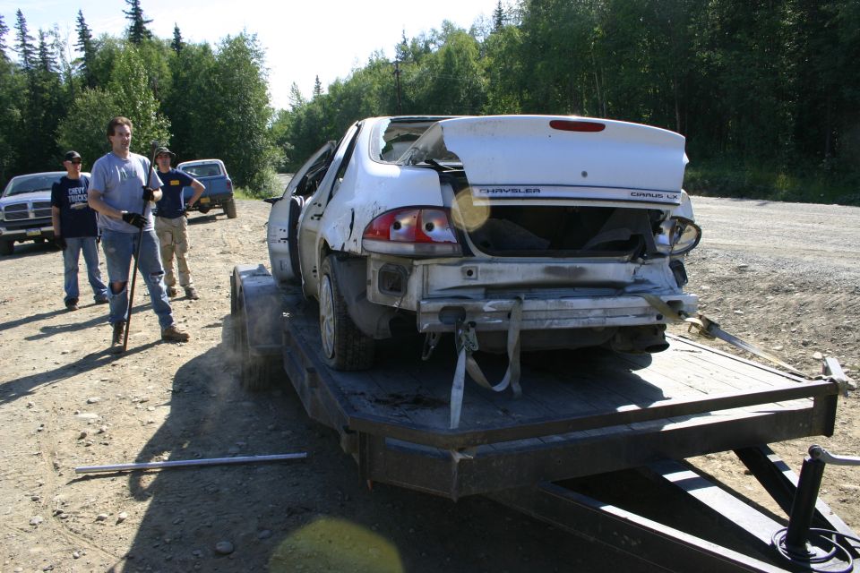 White sedan with rear damage on a trailer, people standing nearby. Chrysler Cirrus LX badge visible