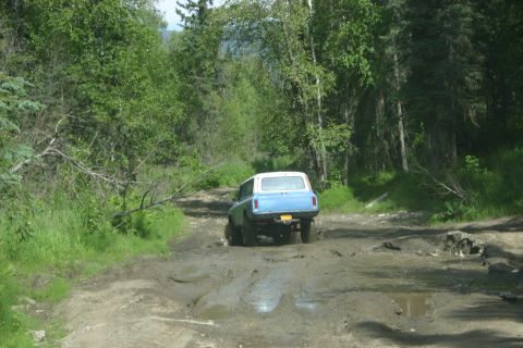 2003 Jeep TJ - A blue SUV navigates a challenging muddy track surrounded by dense green foliage.
