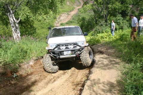 2003 Jeep TJ - A white off-road vehicle with oversized tires navigates a challenging muddy track with onlookers in the background.
