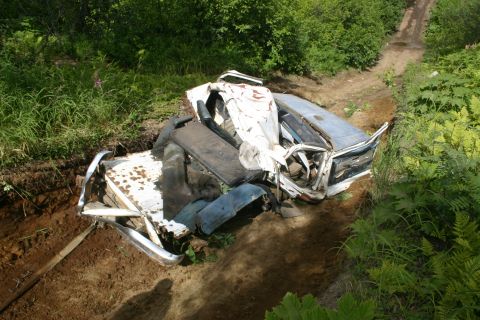 2003 Jeep TJ - A severely damaged white and blue car lies off a dirt road, surrounded by vegetation.