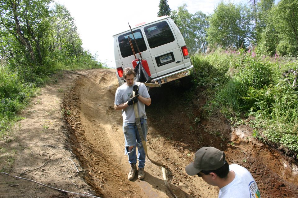 White van partially stuck on a steep dirt incline, with two men nearby. One man holds a tow strap