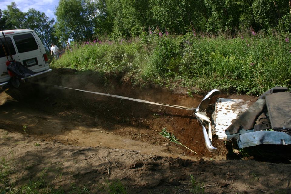 White van pulling a partially buried blue vehicle from a dirt ditch with a tow strap