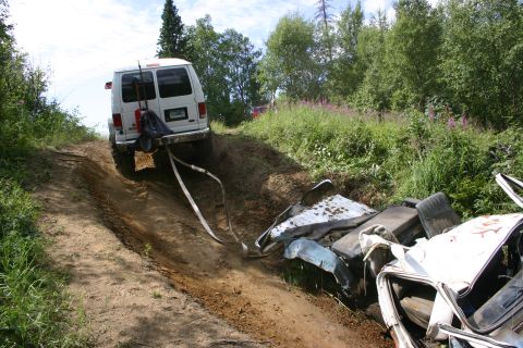 2003 Jeep TJ - A white van is winching a wrecked car from a ditch on a dirt road surrounded by trees and vegetation.