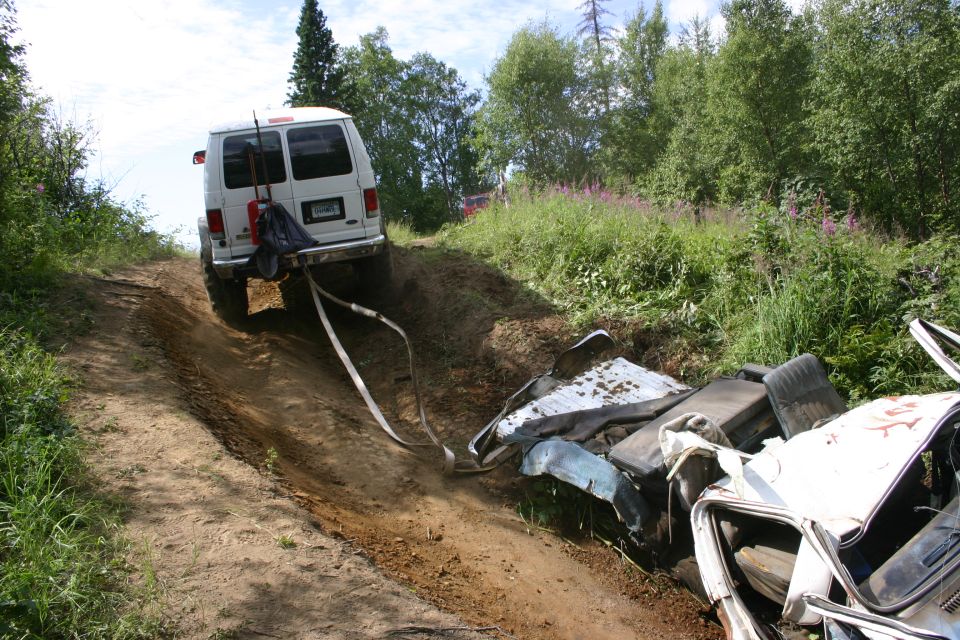 White van on a dirt track pulling a heavily damaged white car off the side of the road
