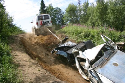 2003 Jeep TJ - A white van tows a heavily damaged car up a steep, dusty incline surrounded by trees and vegetation.